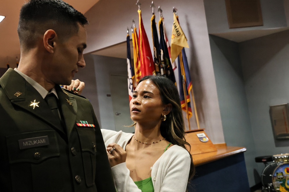 A military officer named Mizukami, wearing a U.S. Army dress green uniform, is having insignia or a pin adjusted on his shoulder by a young woman in a white cardigan and green top. The setting appears to be an official military ceremony room with multiple unit and organizational flags displayed in the background. The interaction suggests a promotion or commissioning ceremony where a family member or significant other is pinning new rank insignia.