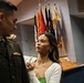 A military officer named Mizukami, wearing a U.S. Army dress green uniform, is having insignia or a pin adjusted on his shoulder by a young woman in a white cardigan and green top. The setting appears to be an official military ceremony room with multiple unit and organizational flags displayed in the background. The interaction suggests a promotion or commissioning ceremony where a family member or significant other is pinning new rank insignia.
