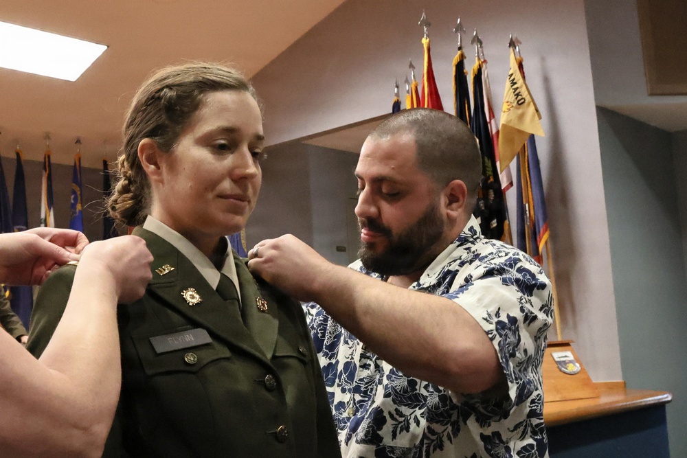 A military promotion or commissioning ceremony is taking place indoors. A woman in a green U.S. Army dress uniform is having insignia or rank pins attached to her collar by a bearded man in a floral shirt, while another person's hands are visible pinning the opposite side. Multiple military and unit flags are displayed in the background.