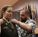 A military promotion or commissioning ceremony is taking place indoors. A woman in a green U.S. Army dress uniform is having insignia or rank pins attached to her collar by a bearded man in a floral shirt, while another person's hands are visible pinning the opposite side. Multiple military and unit flags are displayed in the background.