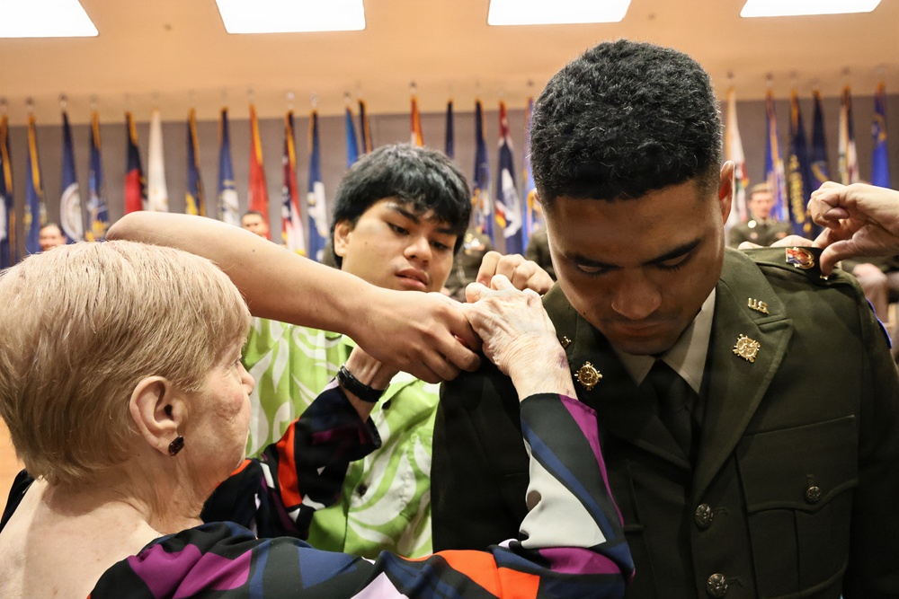 A military promotion or commissioning ceremony is taking place indoors, where an older woman and a younger civilian are pinning insignia or rank devices onto a U.S. Army soldier's dress green uniform. The soldier is looking downward as the pinning occurs, a traditional part of military promotion ceremonies. Multiple state and organizational flags are displayed in the background, indicating a formal military venue.