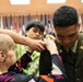 A military promotion or commissioning ceremony is taking place indoors, where an older woman and a younger civilian are pinning insignia or rank devices onto a U.S. Army soldier's dress green uniform. The soldier is looking downward as the pinning occurs, a traditional part of military promotion ceremonies. Multiple state and organizational flags are displayed in the background, indicating a formal military venue.