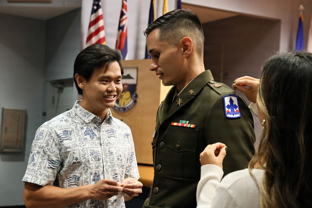 A military promotion or commissioning ceremony is taking place indoors. A soldier in Army dress greens is having rank insignia or pins affixed to his uniform by a woman, while another man in a Hawaiian-style shirt smiles and watches. Multiple flags are visible in the background, indicating an official military setting.