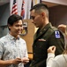 A military promotion or commissioning ceremony is taking place indoors. A soldier in Army dress greens is having rank insignia or pins affixed to his uniform by a woman, while another man in a Hawaiian-style shirt smiles and watches. Multiple flags are visible in the background, indicating an official military setting.