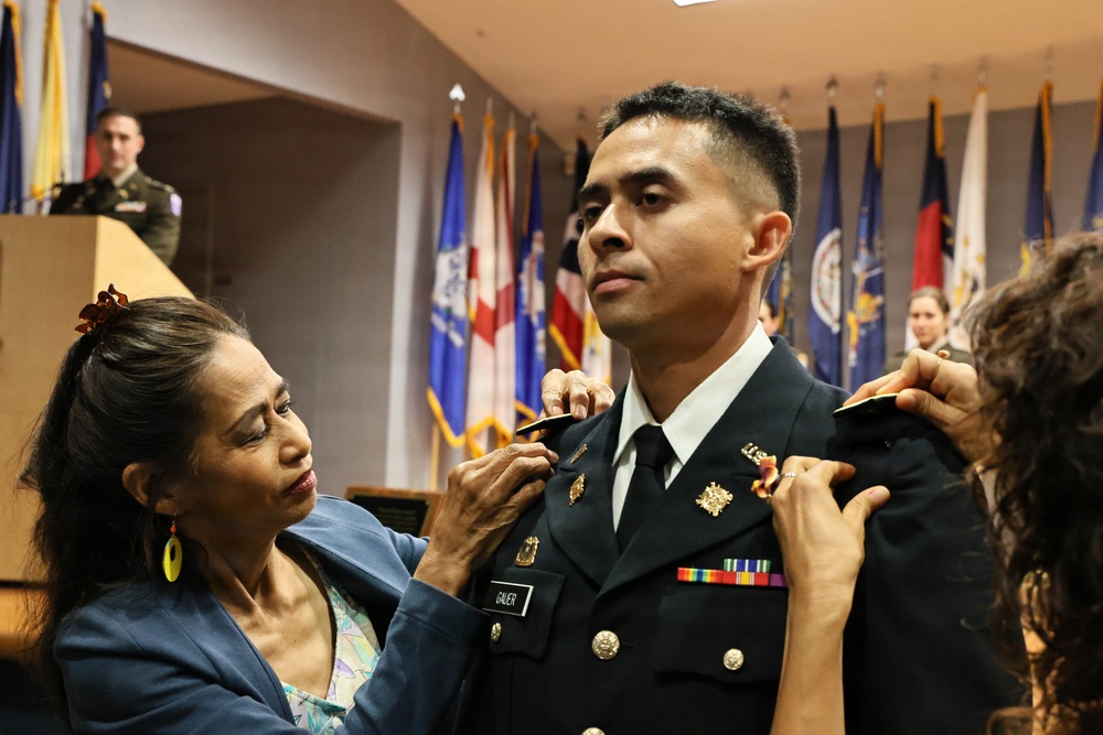 A military commissioning or promotion ceremony is taking place indoors. A woman, likely a family member, is pinning insignia or rank onto the uniform of a male soldier whose name tag reads 'GAUER'. A second person's hands are visible on the soldier's right shoulder, also assisting with the pinning ceremony.