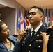 A military commissioning or promotion ceremony is taking place indoors. A woman, likely a family member, is pinning insignia or rank onto the uniform of a male soldier whose name tag reads 'GAUER'. A second person's hands are visible on the soldier's right shoulder, also assisting with the pinning ceremony.