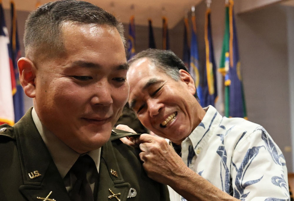 A U.S. Army officer in dress green uniform is having an insignia or rank device pinned onto his shoulder by an older man in a Hawaiian-style shirt, who is smiling broadly. The setting appears to be a formal military promotion or commissioning ceremony, indicated by the multiple flags displayed in the background. The moment captures what appears to be a proud family member participating in the traditional 'pinning' ritual.