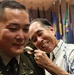 A U.S. Army officer in dress green uniform is having an insignia or rank device pinned onto his shoulder by an older man in a Hawaiian-style shirt, who is smiling broadly. The setting appears to be a formal military promotion or commissioning ceremony, indicated by the multiple flags displayed in the background. The moment captures what appears to be a proud family member participating in the traditional 'pinning' ritual.