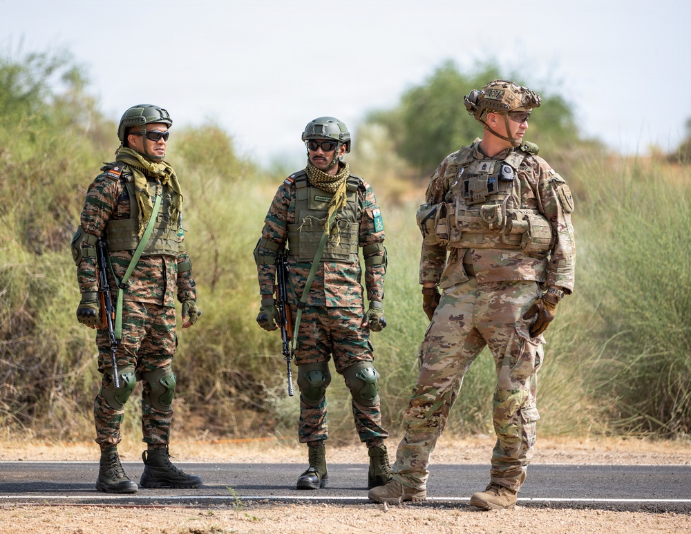 Three armed military personnel are standing on a paved road in an arid, scrubland environment. Two soldiers on the left wear matching camouflage uniforms with green helmets and carry rifles, consistent with Indian Army attire, while the soldier on the right wears MultiCam (OCP) pattern uniform with a more advanced helmet setup, consistent with U.S. Army gear. The scene appears to depict a joint military exercise or patrol.