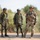 U.S., Indian Soldiers coordinate during Yudh Abhyas 2026 Three armed military personnel are standing on a paved road in an arid, scrubland environment. Two soldiers on the left wear matching camouflage uniforms with green helmets and carry rifles, consistent with Indian Army attire, while the soldier on the right wears MultiCam (OCP) pattern uniform with a more advanced helmet setup, consistent with U.S. Army gear. The scene appears to depict a joint military exercise or patrol.