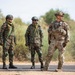 Three armed military personnel are standing on a paved road in an arid, scrubland environment. Two soldiers on the left wear matching camouflage uniforms with green helmets and carry rifles, consistent with Indian Army attire, while the soldier on the right wears MultiCam (OCP) pattern uniform with a more advanced helmet setup, consistent with U.S. Army gear. The scene appears to depict a joint military exercise or patrol.