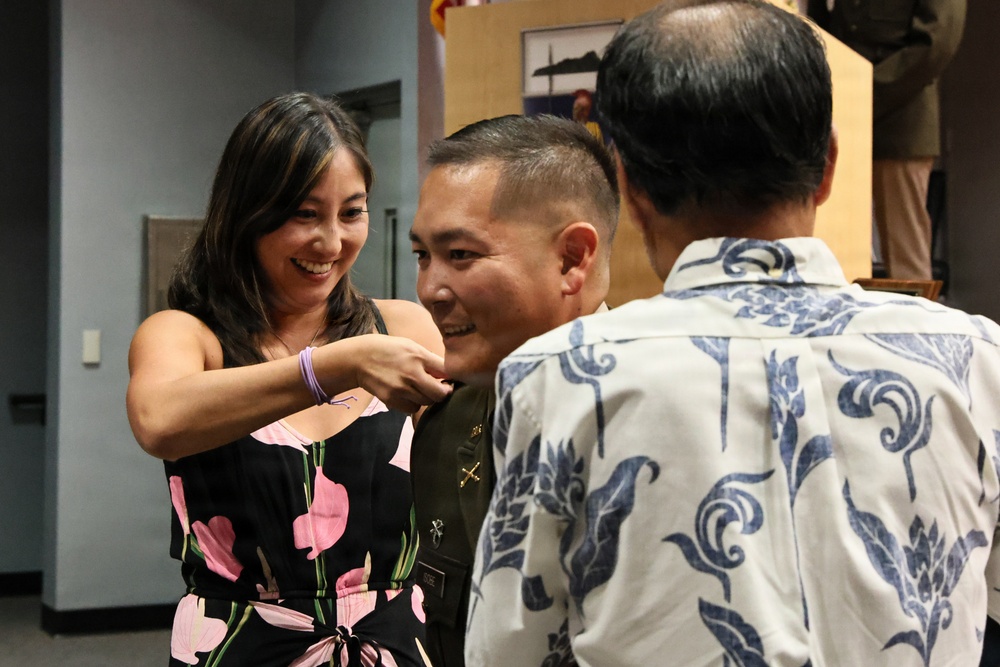A military promotion or pinning ceremony is taking place indoors. A smiling woman in a floral dress is pinning a new insignia or rank onto a uniformed military service member, while a third person in a Hawaiian-style shirt looks on. The atmosphere appears celebratory and joyful.