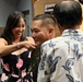 A military promotion or pinning ceremony is taking place indoors. A smiling woman in a floral dress is pinning a new insignia or rank onto a uniformed military service member, while a third person in a Hawaiian-style shirt looks on. The atmosphere appears celebratory and joyful.
