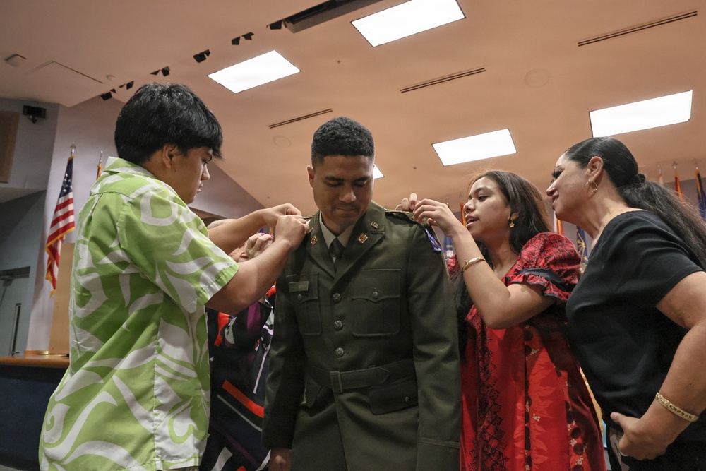 A military promotion or commissioning ceremony is taking place indoors, where family members are pinning new rank insignia onto a soldier's Army Service Uniform. The central figure, wearing an olive green Army dress uniform, stands still while at least three individuals assist in attaching insignia to his collar and shoulders. An American flag and additional flags are visible in the background, consistent with an official military ceremony setting.