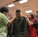 A military promotion or commissioning ceremony is taking place indoors, where family members are pinning new rank insignia onto a soldier's Army Service Uniform. The central figure, wearing an olive green Army dress uniform, stands still while at least three individuals assist in attaching insignia to his collar and shoulders. An American flag and additional flags are visible in the background, consistent with an official military ceremony setting.
