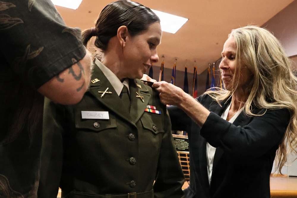 A military promotion or commissioning ceremony is taking place indoors. A woman in a U.S. Army dress green uniform with the name tag 'CAUSEY' is having insignia or rank pinned onto her collar by a civilian woman in a black blazer. A third person, partially visible on the left with a visible tattoo, is also participating in the pinning ceremony.