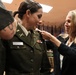A military promotion or commissioning ceremony is taking place indoors. A woman in a U.S. Army dress green uniform with the name tag 'CAUSEY' is having insignia or rank pinned onto her collar by a civilian woman in a black blazer. A third person, partially visible on the left with a visible tattoo, is also participating in the pinning ceremony.