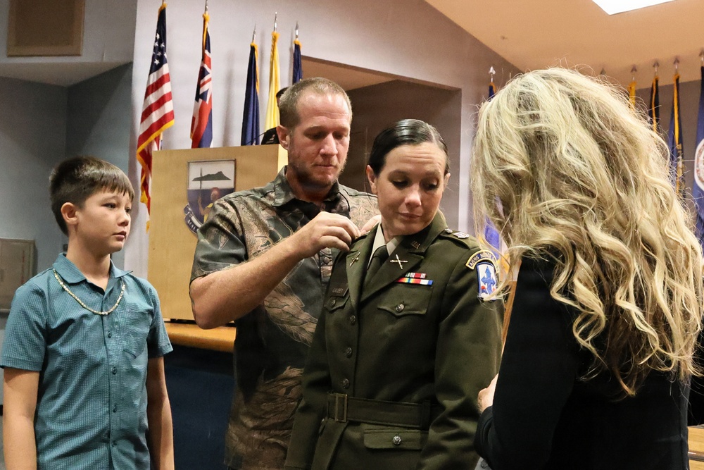 A military promotion or pinning ceremony is taking place indoors, likely at a U.S. Army installation. A woman in Army green service uniform is having insignia pinned onto her collar by a man in civilian clothing, while a blonde woman assists on the other side and a young boy watches. Multiple U.S. and state flags are visible in the background.