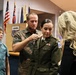 A military promotion or pinning ceremony is taking place indoors, likely at a U.S. Army installation. A woman in Army green service uniform is having insignia pinned onto her collar by a man in civilian clothing, while a blonde woman assists on the other side and a young boy watches. Multiple U.S. and state flags are visible in the background.