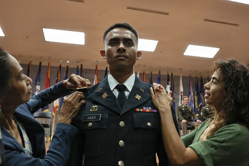 A U.S. Army soldier named GAUER is having rank insignia pinned onto his dress uniform by two women, likely family members, during a formal military commissioning or promotion ceremony. The soldier stands at attention in Army Service Uniform (ASU) while the women attach pins to each shoulder. The ceremony is taking place in an official venue decorated with multiple state and military flags.