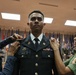 A U.S. Army soldier named GAUER is having rank insignia pinned onto his dress uniform by two women, likely family members, during a formal military commissioning or promotion ceremony. The soldier stands at attention in Army Service Uniform (ASU) while the women attach pins to each shoulder. The ceremony is taking place in an official venue decorated with multiple state and military flags.