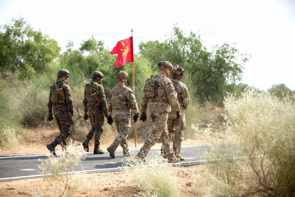 A group of five military personnel are walking along a paved road in an arid, scrubland environment. The group includes soldiers in two distinct camouflage patterns — US Army MultiCam (OCP) and a green tiger-stripe or foreign pattern — suggesting a joint or multinational military exercise. A red guidon flag with yellow insignia, bearing what appears to be the number '8' and a unit emblem, is being carried by one of the soldiers.