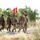 U.S., Indian Soldiers move together during Yudh Abhyas 2026 A group of five military personnel are walking along a paved road in an arid, scrubland environment. The group includes soldiers in two distinct camouflage patterns — US Army MultiCam (OCP) and a green tiger-stripe or foreign pattern — suggesting a joint or multinational military exercise. A red guidon flag with yellow insignia, bearing what appears to be the number '8' and a unit emblem, is being carried by one of the soldiers.