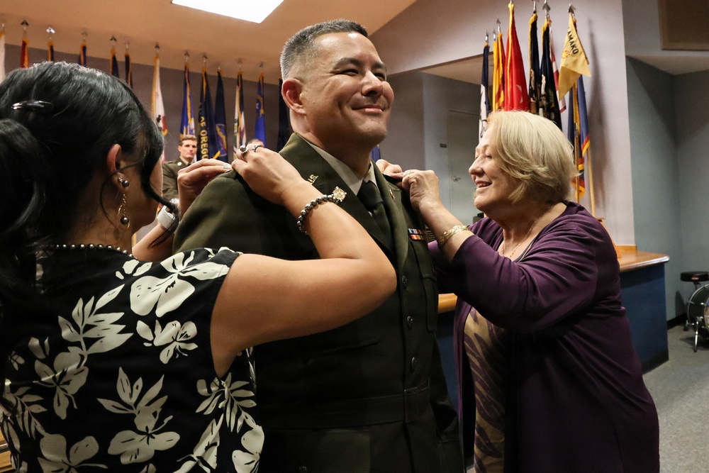 A military promotion ceremony is taking place indoors, where a uniformed soldier is having rank insignia pinned onto his collar by two women, likely family members. The soldier is smiling broadly, suggesting this is a celebratory milestone event. Multiple military flags are displayed in the background, consistent with an official military setting.