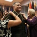A military promotion ceremony is taking place indoors, where a uniformed soldier is having rank insignia pinned onto his collar by two women, likely family members. The soldier is smiling broadly, suggesting this is a celebratory milestone event. Multiple military flags are displayed in the background, consistent with an official military setting.