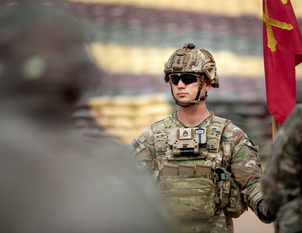 A U.S. Army soldier in full combat gear stands at attention during what appears to be a formal military ceremony or formation. The soldier wears Operational Camouflage Pattern (OCP) uniform with a tactical helmet, body armor, and rank insignia visible on the chest plate indicating the rank of Staff Sergeant. A red guidon flag with gold crossed cannons (Field Artillery branch insignia) is visible in the background.