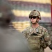 A U.S. Army soldier in full combat gear stands at attention during what appears to be a formal military ceremony or formation. The soldier wears Operational Camouflage Pattern (OCP) uniform with a tactical helmet, body armor, and rank insignia visible on the chest plate indicating the rank of Staff Sergeant. A red guidon flag with gold crossed cannons (Field Artillery branch insignia) is visible in the background.