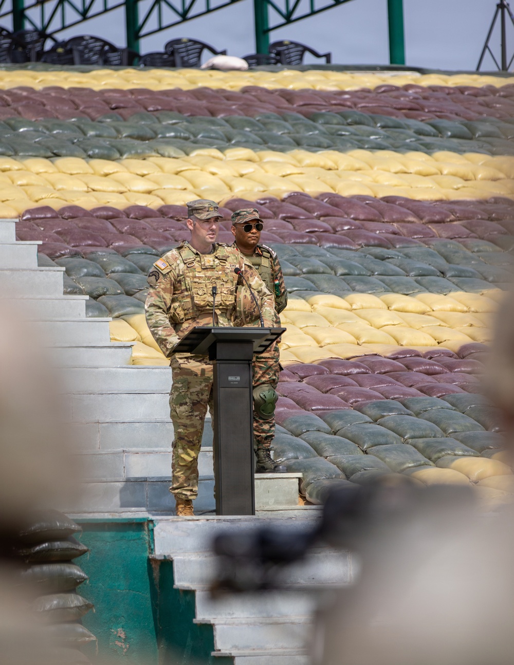 Two military personnel are standing at a podium in what appears to be an outdoor ceremonial or briefing setting. One soldier in US Army MultiCam/OCP uniform with a tactical vest is speaking at the podium, while a second soldier in a different military uniform stands behind. The background features a large display of sandbags arranged in alternating colored rows of yellow, maroon/brown, and green/gray.