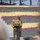 U.S. Army Leader addresses Yudh Abhyas 2026 formation Two military personnel are standing at a podium in what appears to be an outdoor ceremonial or briefing setting. One soldier in US Army MultiCam/OCP uniform with a tactical vest is speaking at the podium, while a second soldier in a different military uniform stands behind. The background features a large display of sandbags arranged in alternating colored rows of yellow, maroon/brown, and green/gray.