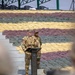 Two military personnel are standing at a podium in what appears to be an outdoor ceremonial or briefing setting. One soldier in US Army MultiCam/OCP uniform with a tactical vest is speaking at the podium, while a second soldier in a different military uniform stands behind. The background features a large display of sandbags arranged in alternating colored rows of yellow, maroon/brown, and green/gray.