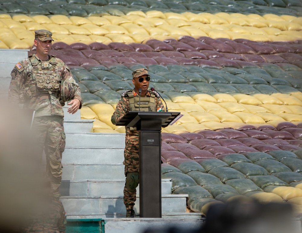 Two military personnel are present at an outdoor podium, with one officer in Indian Army camouflage uniform speaking at a lectern while a US Army soldier stands nearby. The background features a large arrangement of sandbags in multiple colors — yellow, maroon/red, and green — arranged in horizontal rows, likely forming a decorative or symbolic display. The setting appears to be a formal military ceremony or joint exercise event.