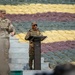 Two military personnel are present at an outdoor podium, with one officer in Indian Army camouflage uniform speaking at a lectern while a US Army soldier stands nearby. The background features a large arrangement of sandbags in multiple colors — yellow, maroon/red, and green — arranged in horizontal rows, likely forming a decorative or symbolic display. The setting appears to be a formal military ceremony or joint exercise event.