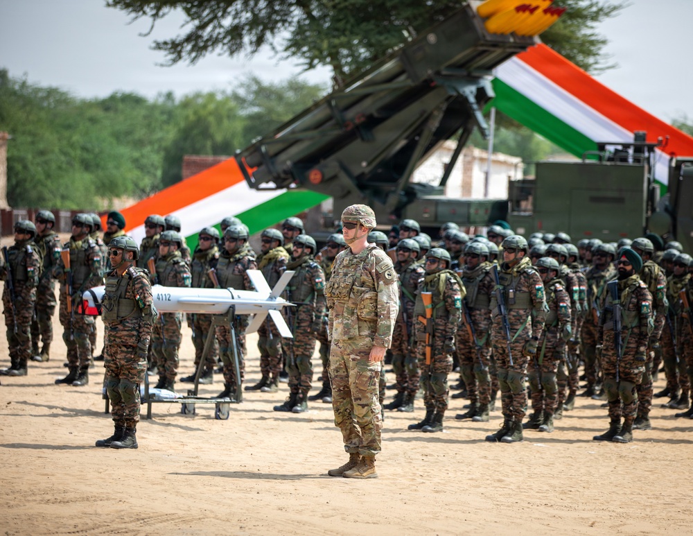 A military parade or joint exercise ceremony featuring soldiers from what appears to be India and the United States, standing in formation on sandy terrain. The Indian tricolor flag is prominently displayed in the background alongside what appears to be a missile launcher system. A small fixed-wing UAV/drone (numbered 4112) is displayed on a stand in the foreground.