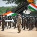 A military parade or joint exercise ceremony featuring soldiers from what appears to be India and the United States, standing in formation on sandy terrain. The Indian tricolor flag is prominently displayed in the background alongside what appears to be a missile launcher system. A small fixed-wing UAV/drone (numbered 4112) is displayed on a stand in the foreground.