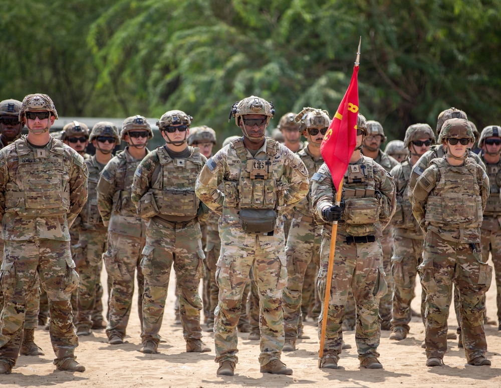 A formation of U.S. Army soldiers standing at ease in what appears to be an outdoor ceremony or formation on sandy terrain. The soldiers are wearing Operational Camouflage Pattern (OCP) uniforms with combat gear, helmets, and protective eyewear. A guidon bearer is visible holding a red unit flag with yellow markings.