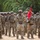 U.S. Soldiers stand in formation during Yudh Abhyas 2026 A formation of U.S. Army soldiers standing at ease in what appears to be an outdoor ceremony or formation on sandy terrain. The soldiers are wearing Operational Camouflage Pattern (OCP) uniforms with combat gear, helmets, and protective eyewear. A guidon bearer is visible holding a red unit flag with yellow markings.