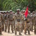 A formation of U.S. Army soldiers standing at ease in what appears to be an outdoor ceremony or formation on sandy terrain. The soldiers are wearing Operational Camouflage Pattern (OCP) uniforms with combat gear, helmets, and protective eyewear. A guidon bearer is visible holding a red unit flag with yellow markings.