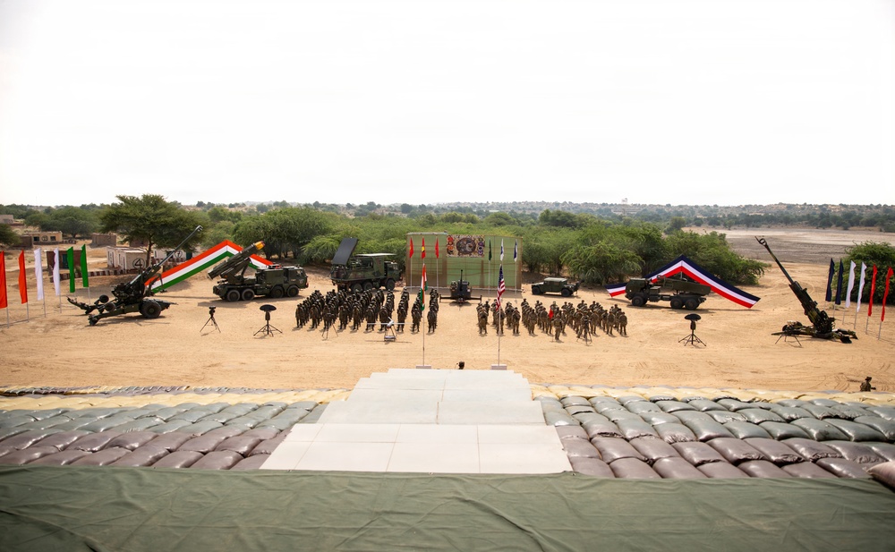 A formal military ceremony or parade is taking place in an arid, desert-like outdoor setting. Multiple nations appear to be represented, with visible flags including those of India, the United States, and at least one other nation, along with artillery pieces and military vehicles displayed on either side. Troops are assembled in formation in front of a central stage or podium area.