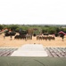 A formal military ceremony or parade is taking place in an arid, desert-like outdoor setting. Multiple nations appear to be represented, with visible flags including those of India, the United States, and at least one other nation, along with artillery pieces and military vehicles displayed on either side. Troops are assembled in formation in front of a central stage or podium area.