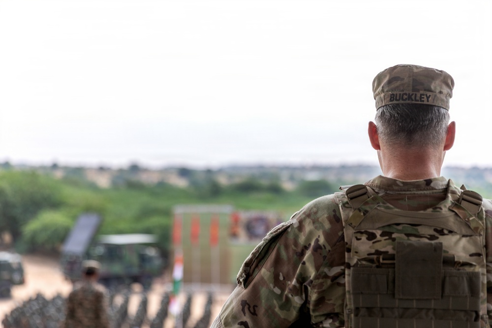 A military personnel member identified as 'BUCKLEY' on their patrol cap is photographed from behind, wearing Operational Camouflage Pattern (OCP) uniform and tactical vest. The individual appears to be observing a military training area or operational site with vehicles, other personnel, and range markers visible in the blurred background. The image uses a shallow depth of field, keeping the foreground subject sharp while the background remains out of focus.