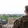 U.S. Army leader observes Yudh Abhyas 2026 Opening Ceremony A military personnel member identified as 'BUCKLEY' on their patrol cap is photographed from behind, wearing Operational Camouflage Pattern (OCP) uniform and tactical vest. The individual appears to be observing a military training area or operational site with vehicles, other personnel, and range markers visible in the blurred background. The image uses a shallow depth of field, keeping the foreground subject sharp while the background remains out of focus.
