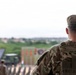 A military personnel member identified as 'BUCKLEY' on their patrol cap is photographed from behind, wearing Operational Camouflage Pattern (OCP) uniform and tactical vest. The individual appears to be observing a military training area or operational site with vehicles, other personnel, and range markers visible in the blurred background. The image uses a shallow depth of field, keeping the foreground subject sharp while the background remains out of focus.
