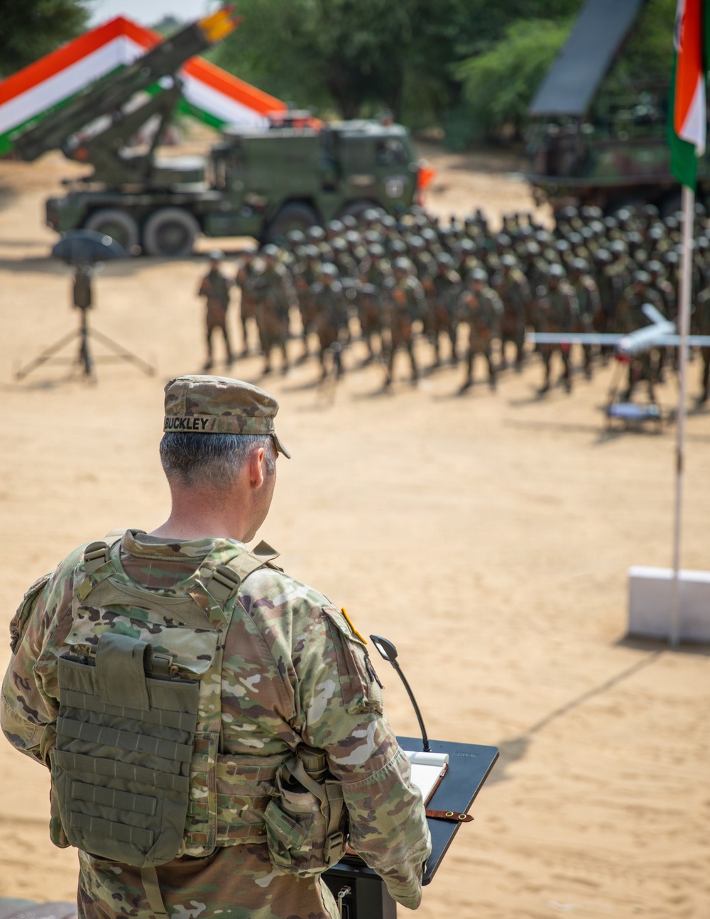 A U.S. Army soldier named Buckley stands at a podium addressing a large formation of troops during what appears to be a joint military exercise or ceremony. Indian national flags (tricolor: saffron, white, and green) are prominently displayed alongside military vehicles including what appears to be a multi-barrel rocket launcher system. The setting is an arid, sandy outdoor area with trees in the background.