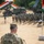 U.S. Army leader addresses Soldiers at Yudh Abhyas 2026 A U.S. Army soldier named Buckley stands at a podium addressing a large formation of troops during what appears to be a joint military exercise or ceremony. Indian national flags (tricolor: saffron, white, and green) are prominently displayed alongside military vehicles including what appears to be a multi-barrel rocket launcher system. The setting is an arid, sandy outdoor area with trees in the background.
