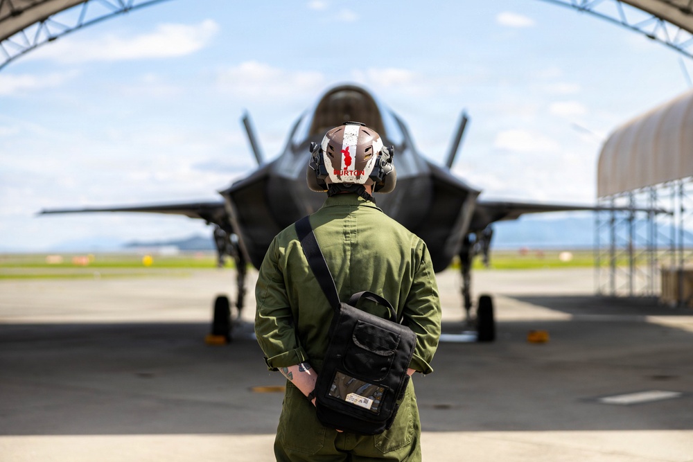 A military personnel member wearing a green flight suit and a white helmet with 'BURTON' written on it stands facing an F-35 Lightning II stealth fighter jet parked under a hangar arch. The individual carries a black shoulder bag and has visible tattoos on their arm. The scene appears to be on an active military airfield under partly cloudy skies.
