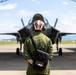 A military personnel member wearing a green flight suit and a white helmet with 'BURTON' written on it stands facing an F-35 Lightning II stealth fighter jet parked under a hangar arch. The individual carries a black shoulder bag and has visible tattoos on their arm. The scene appears to be on an active military airfield under partly cloudy skies.